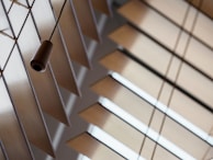 A close-up view of horizontal wooden blinds with light coming through the gaps, creating geometric patterns. A dark brown cord with a wooden pull hangs in the foreground, adding depth to the composition.