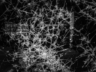 Intricate web-like patterns of shattered glass spread across the surface, with cracks radiating outward from multiple points of impact. The background is dark, contrasting sharply with the reflections and highlights on the jagged edges of the glass.