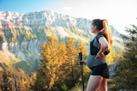 A pregnant woman stands in a mountainous landscape, holding trekking poles and looking at the scenery. She has long hair tied back and is wearing outdoor athletic gear. The background features lush trees and towering cliffs with a clear blue sky above.