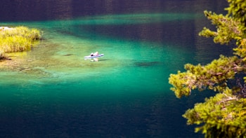A serene landscape featuring a small boat with a person reclining, surrounded by calm, turquoise waters. Lush green trees and grasses line the shoreline, enhancing the natural beauty of the scene.