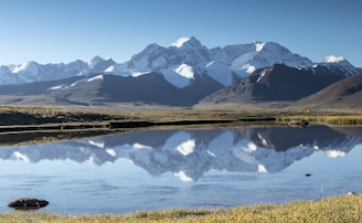 Snow-capped mountains with a serene lake reflecting the clear blue sky and surrounding nature.