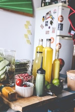 The image depicts a kitchen scene with various ingredients and items on a wooden table. There are bottles filled with yellow liquid, possibly juice or sauce, a green squeeze bottle, limes, red tomatoes, and red berries in a white bowl. In the background, there's a refrigerator covered in magnets and photos, with additional kitchen items such as a glass jar with corn.