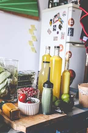 The image depicts a kitchen scene with various ingredients and items on a wooden table. There are bottles filled with yellow liquid, possibly juice or sauce, a green squeeze bottle, limes, red tomatoes, and red berries in a white bowl. In the background, there's a refrigerator covered in magnets and photos, with additional kitchen items such as a glass jar with corn.