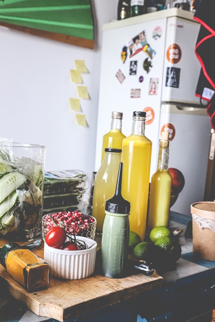 The image depicts a kitchen scene with various ingredients and items on a wooden table. There are bottles filled with yellow liquid, possibly juice or sauce, a green squeeze bottle, limes, red tomatoes, and red berries in a white bowl. In the background, there's a refrigerator covered in magnets and photos, with additional kitchen items such as a glass jar with corn.