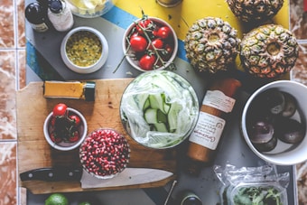 An assortment of fresh ingredients is laid out on a kitchen surface, including pineapples, cherry tomatoes, cucumbers, pomegranate seeds, passion fruit, and spices. A wooden cutting board holds some of the ingredients along with a knife. Nearby, there are bottles and other kitchen items.