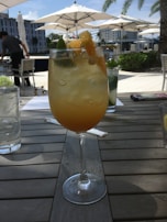 A cocktail glass filled with an orange drink garnished with lime and orange slices sits on a wooden table. In the background, outdoor patio furniture and white umbrellas provide shade, with palm trees and a building visible under a clear blue sky.