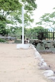 A cat sits near the base of a white signpost in a tranquil outdoor setting. The ground is covered in sand and bordered by neatly arranged stones. In the background, green trees and a rustic wooden fence create a natural, peaceful environment.