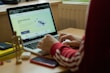 A person in a red sweatshirt is typing on a laptop, which displays a webpage about 'Simple Author Box Pro' on the screen. The setting appears to be a study or home office, with a desk that holds a smartphone, wooden figure, and stack of books for decoration. Soft natural light comes from a nearby window.