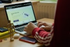 A person in a red sweatshirt is typing on a laptop, which displays a webpage about 'Simple Author Box Pro' on the screen. The setting appears to be a study or home office, with a desk that holds a smartphone, wooden figure, and stack of books for decoration. Soft natural light comes from a nearby window.