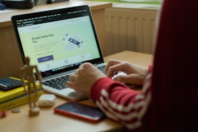 A person in a red sweatshirt is typing on a laptop, which displays a webpage about 'Simple Author Box Pro' on the screen. The setting appears to be a study or home office, with a desk that holds a smartphone, wooden figure, and stack of books for decoration. Soft natural light comes from a nearby window.