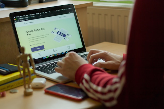 A person in a red sweatshirt is typing on a laptop, which displays a webpage about 'Simple Author Box Pro' on the screen. The setting appears to be a study or home office, with a desk that holds a smartphone, wooden figure, and stack of books for decoration. Soft natural light comes from a nearby window.