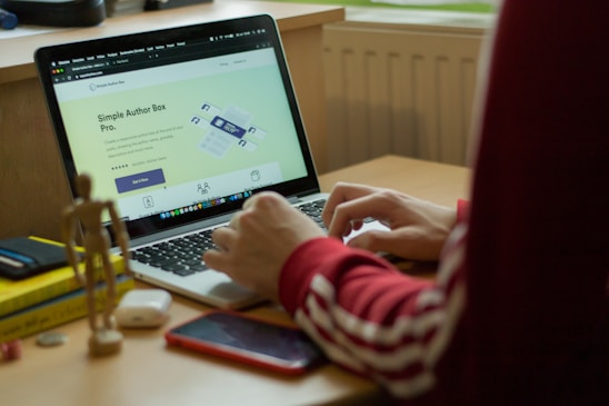 A person in a red sweatshirt is typing on a laptop, which displays a webpage about 'Simple Author Box Pro' on the screen. The setting appears to be a study or home office, with a desk that holds a smartphone, wooden figure, and stack of books for decoration. Soft natural light comes from a nearby window.