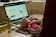 A person in a red sweatshirt is typing on a laptop, which displays a webpage about 'Simple Author Box Pro' on the screen. The setting appears to be a study or home office, with a desk that holds a smartphone, wooden figure, and stack of books for decoration. Soft natural light comes from a nearby window.