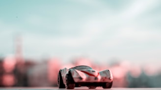 A small, toy car is positioned on a flat surface with a blurred, pastel-colored background. The toy car is detailed with markings and has an aerodynamic design. The scene is softly lit, creating a dreamy atmosphere.