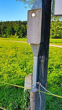A wooden pole with metal fixtures is set in a green field under a clear blue sky. The foreground pole has an electrical box attached, and wires run down it. Lush vegetation surrounds the area, suggesting a rural or countryside setting.