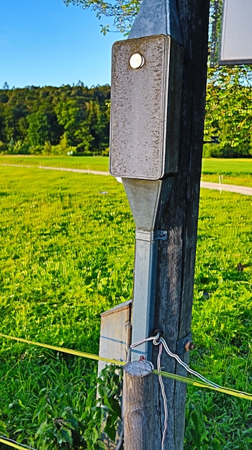 A wooden pole with metal fixtures is set in a green field under a clear blue sky. The foreground pole has an electrical box attached, and wires run down it. Lush vegetation surrounds the area, suggesting a rural or countryside setting.