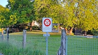 A grassy area with a wire fence is bordered by a line of wooden posts. A sign indicating private property restricts access for cars and motorcycles. Trees with lush green foliage provide shade in the background, and sunlight bathes the scene, highlighting the details in the landscape.