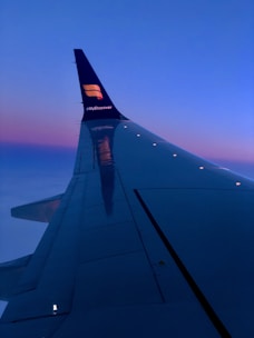 The image features the wing of an airplane viewed from a passenger window, with a vivid gradient in the sky transitioning from a rich blue to a lighter pink hue near the horizon. The airplane's tail displays a logo and a hashtag.