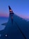 The image features the wing of an airplane viewed from a passenger window, with a vivid gradient in the sky transitioning from a rich blue to a lighter pink hue near the horizon. The airplane's tail displays a logo and a hashtag.