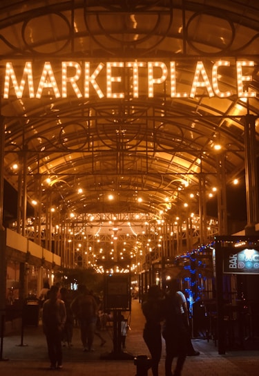 A warmly lit marketplace entrance at night, with illuminated letters overhead spelling 'MARKETPLACE'. Strings of glowing bulbs create a cozy and inviting atmosphere. People walk beneath the canopy, and a bar with a neon sign can be seen on the right.