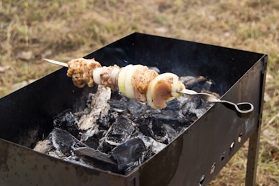 A skewer with pieces of marinated meat and onions is placed on a metal grill over charred coals. The barbecue is situated outdoors on a grassy surface.