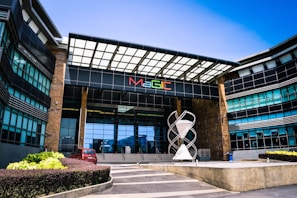 A modern building with a large glass entrance and an overhanging roof structure, featuring a colorful 'MaGIC' sign. In front, there is a distinctive sculpture resembling interlocking shapes and a small red car parked on the left. The building has symmetrical design elements with glass windows and stone accents.