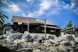 A rustic wooden house with a corrugated iron roof stands in a rural setting. Rocks and rubble are piled in the foreground. The sky is partly cloudy with patches of blue, and palm trees can be seen nearby.