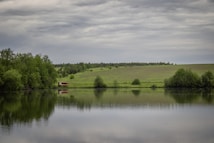 A serene landscape with a reflective lake in the foreground, surrounded by lush green trees on one side and an expansive grassy field on the other. A small wooden structure is visible near the water's edge, under a cloudy sky.