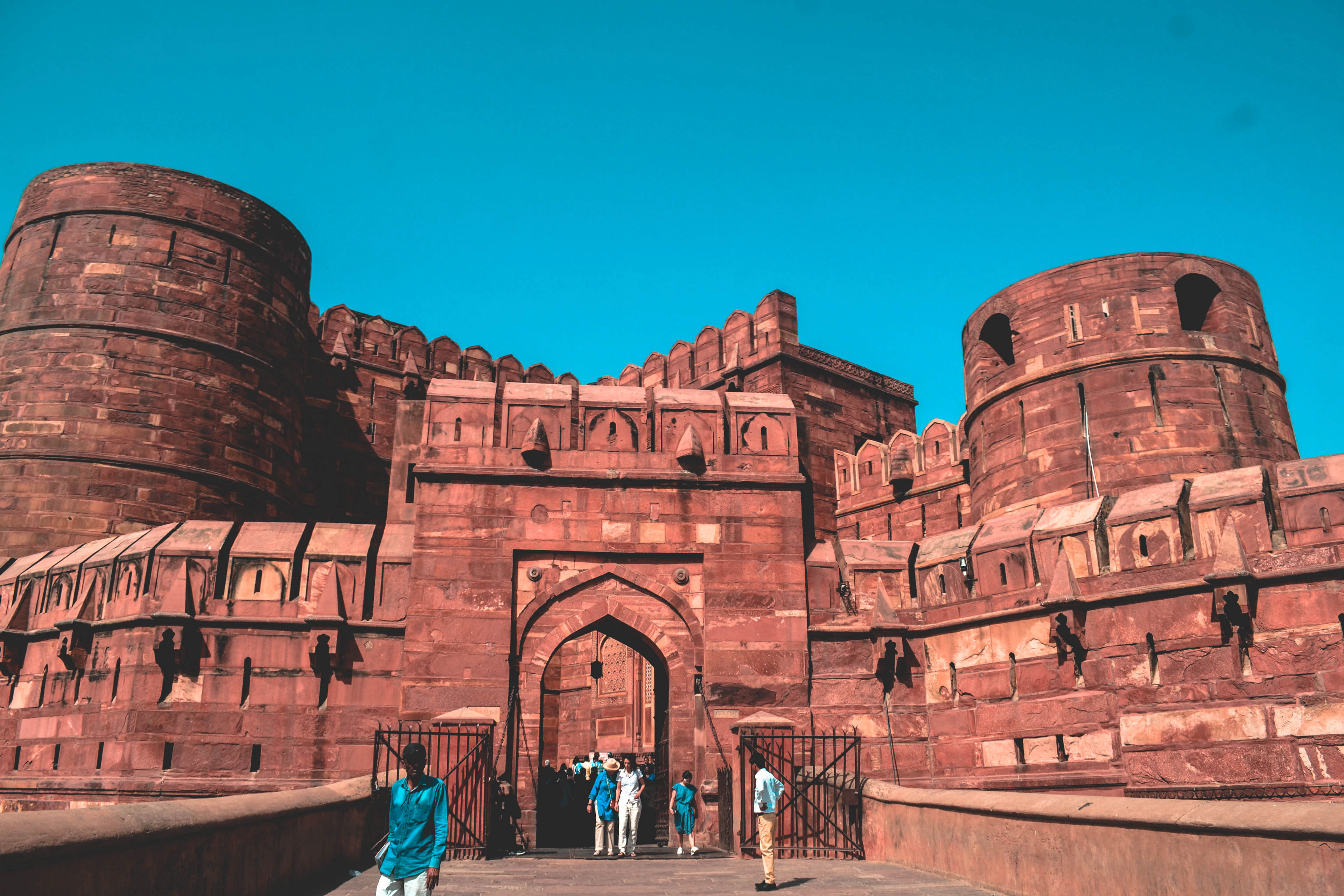 The Amar Singh Gate of Agra Fort: two massive red sandstone bastions flanking an arched entrance, the crenellated walls glowing warm against a deep blue sky as visitors walk up the ramp.