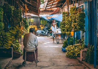 A narrow street market with bunches of bananas hanging from the roof on both sides. There is a person sitting on a stool facing away, and another person is seen in the distance with a bicycle cart. The market has a rustic feel with hanging wooden panels and a scooter parked in the background.
