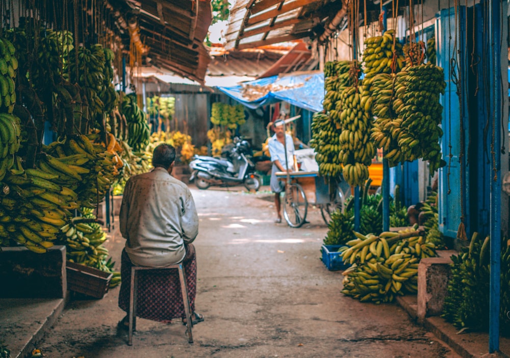 homme en chemise grise assis sur une chaise en bois marron près d’un magasin de fruits de banane pendant la journée