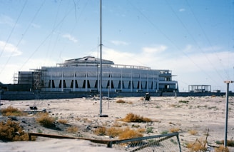 A large, modernist building under construction with a distinctive triangular-patterned facade stands in a barren, sandy landscape. The sky is clear with a few clouds. Scaffolding is visible on one side of the building, and there are construction materials scattered on the ground.