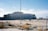 A large, modernist building under construction with a distinctive triangular-patterned facade stands in a barren, sandy landscape. The sky is clear with a few clouds. Scaffolding is visible on one side of the building, and there are construction materials scattered on the ground.