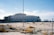 A large, modernist building under construction with a distinctive triangular-patterned facade stands in a barren, sandy landscape. The sky is clear with a few clouds. Scaffolding is visible on one side of the building, and there are construction materials scattered on the ground.