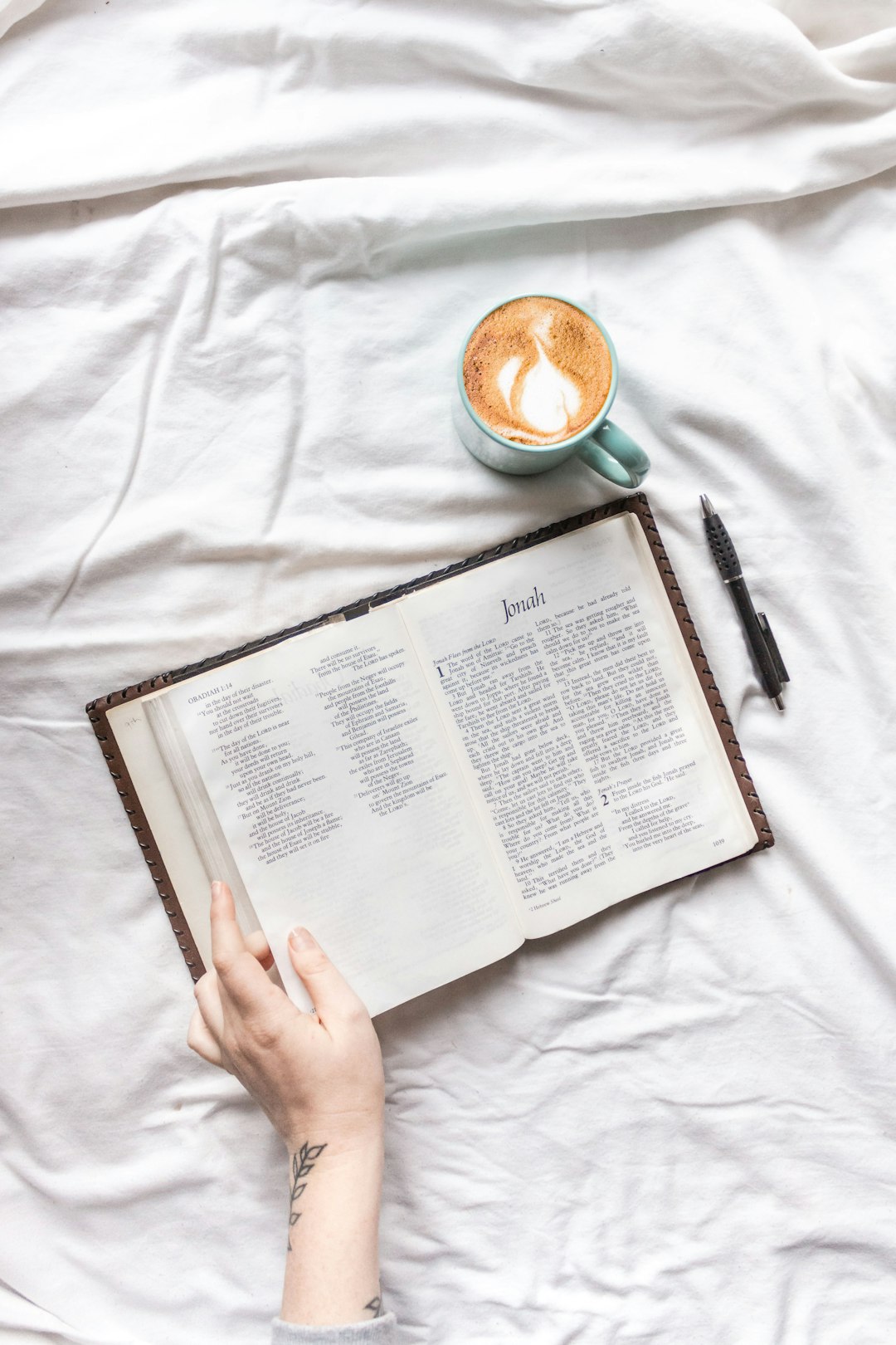 person holding white book page beside black pen and white ceramic mug on white textile