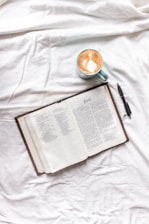 A book is laid open on a white fabric surface next to a cappuccino in a light green cup. Beside the book is a black pen. The book appears to be a Bible open to the Book of Jonah.