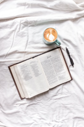 A book is laid open on a white fabric surface next to a cappuccino in a light green cup. Beside the book is a black pen. The book appears to be a Bible open to the Book of Jonah.