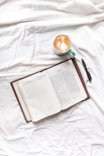 An open book with a leather cover rests on a white fabric surface, displaying text from the book of Acts. Nearby, a cup of cappuccino in a greenish-blue mug with latte art and a black pen are placed on the sheet.