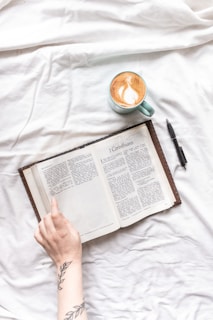 An open book resting on a white fabric surface with a person's hand showing part of an arm tattoo and a pen laying nearby. A light blue cup filled with coffee featuring latte art sits next to the book.