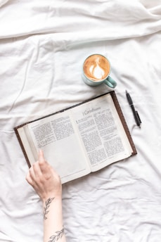 An open book resting on a white fabric surface with a person's hand showing part of an arm tattoo and a pen laying nearby. A light blue cup filled with coffee featuring latte art sits next to the book.