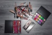 A variety of makeup items are arranged on a wooden surface. There are numerous brushes, lipsticks, and two palettes—one with a wide array of colorful eyeshadows and another with different shades of lip colors. The items are laid out in an organized manner, showcasing the diversity of makeup products.