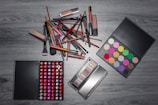 A variety of makeup items are arranged on a wooden surface. There are numerous brushes, lipsticks, and two palettes—one with a wide array of colorful eyeshadows and another with different shades of lip colors. The items are laid out in an organized manner, showcasing the diversity of makeup products.