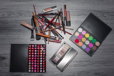 A variety of makeup items are arranged on a wooden surface. There are numerous brushes, lipsticks, and two palettes—one with a wide array of colorful eyeshadows and another with different shades of lip colors. The items are laid out in an organized manner, showcasing the diversity of makeup products.