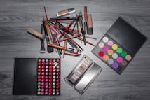 A variety of makeup items are arranged on a wooden surface. There are numerous brushes, lipsticks, and two palettes&mdash;one with a wide array of colorful eyeshadows and another with different shades of lip colors. The items are laid out in an organized manner, showcasing the diversity of makeup products.