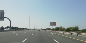 A wide, multi-lane highway stretches into the distance under a clear blue sky. The road is lined with greenery and trees on both sides. There are a few vehicles on the road, including one prominently visible in the distance. Billboards are placed along the side of the highway.