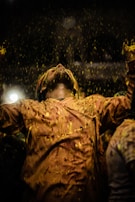 A person is celebrating with arms raised and head tilted back, covered in yellow powder. The background is dark, accentuating the vibrant powder raining down on the individual. The scene captures a moment of joy and festivity.
