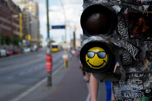 A traffic light with a poster of a yellow smiley face wearing sunglasses is placed on one light, surrounded by taped and torn advertisements. A city street with buildings and a blurred background can be seen.