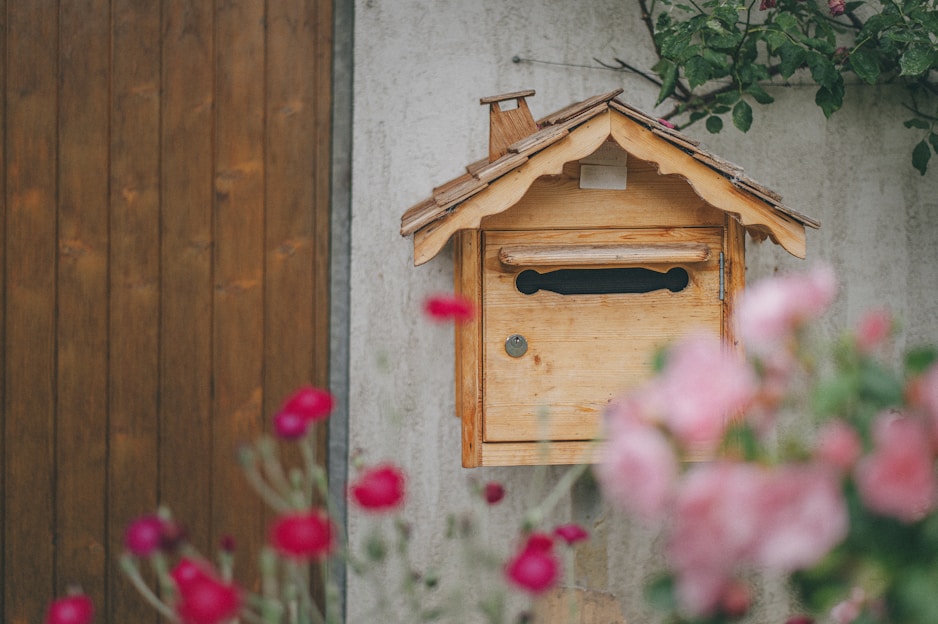 A wooden mailbox designed like a small house is mounted on a wall. The box has a pitched roof and a slot for mail. Pink and red flowers are blurred in the foreground, adding a touch of color. A wooden door and green leaves frame the background.