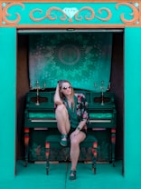 A person wearing sunglasses and casual clothing sits on a chair in front of a teal-colored piano. The setting is framed by an ornately decorated teal and orange arch. Behind the piano, a tapestry with an intricate mandala pattern hangs. Two candle holders adorn the piano, adding a touch of elegance.