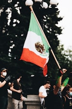 A person in a crowd raises a large Mexican flag with a green, white, and red color scheme, featuring an eagle emblem in the center. The scene shows multiple people, some wearing masks and gloves, possibly indicating a protest or public gathering. The background includes trees and the base of a lamp post.